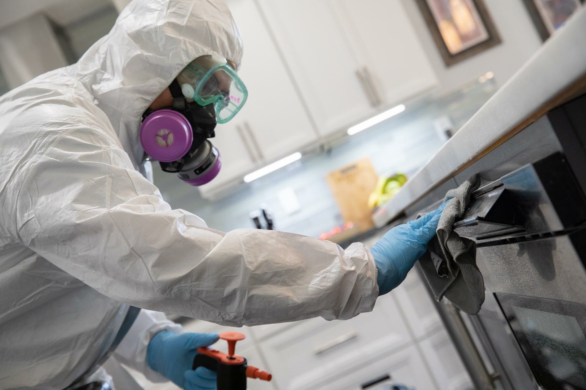 Technician wearing a white biohazard suit, goggles and half face respirator wiping and disinfecting a microwave of COVID-19