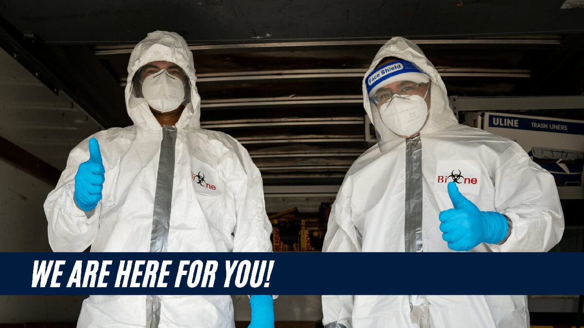 Two Bio-One service technicians dressed in white hazmat suits, N-95 masks and face shield giving a thumbs up.   We Are Here For You is on a title banner on the bottom left of the image.   Bio-One logo is on the left breast of both hazmat suits.